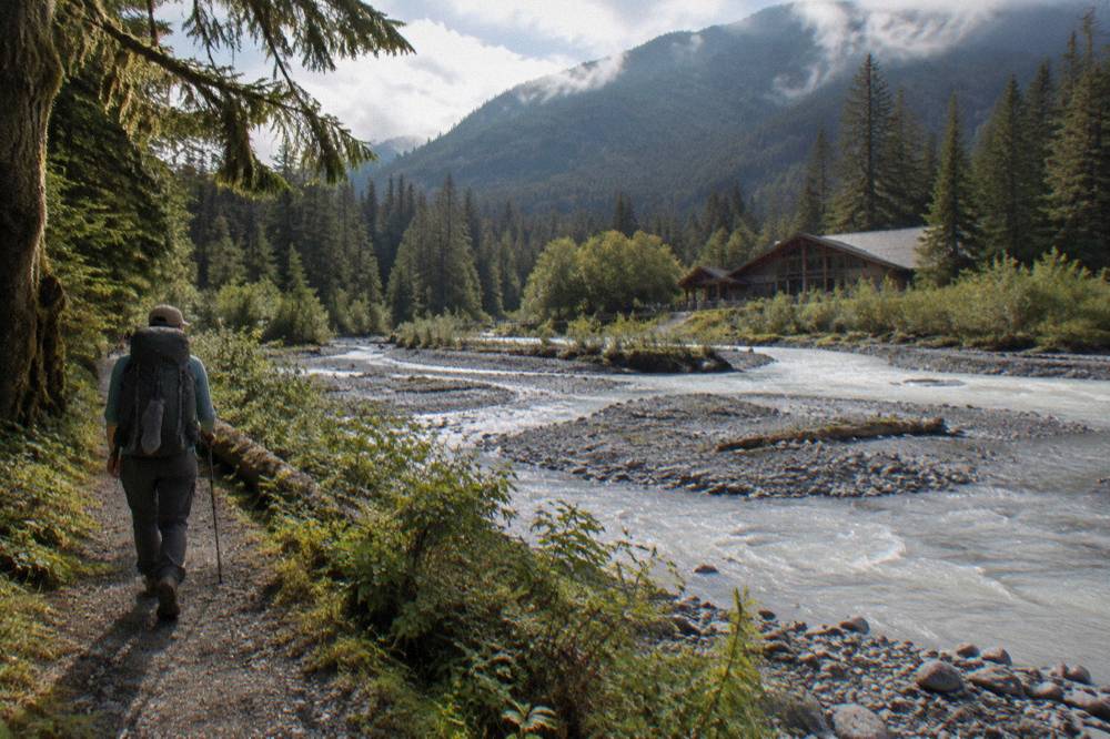 A hiker with a backpack walking a forest path beside a glacial river