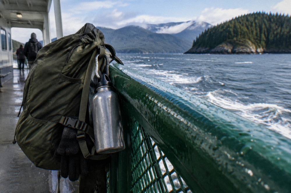 A daypack and water bottle resting on a ferry deck rail with forested islands in the distance