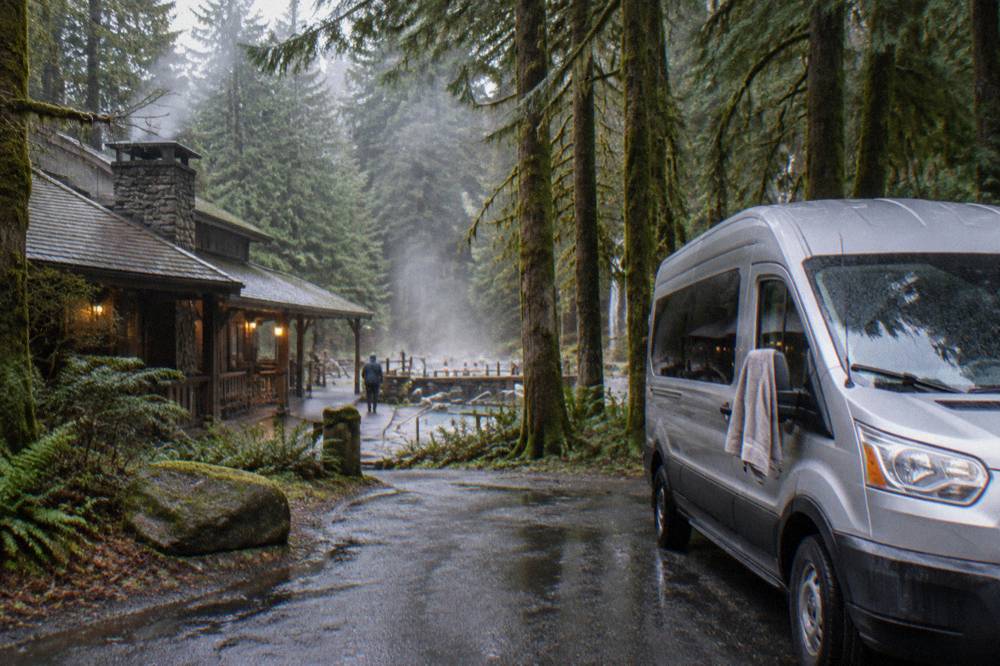 A van parked at a misty rainforest trailhead beside tall mossy trees