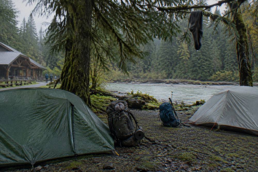 Backpacks and tents arranged at a forest campsite beside a river