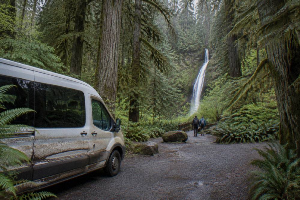 A small tour van parked at a forest trailhead beside tall evergreen trees