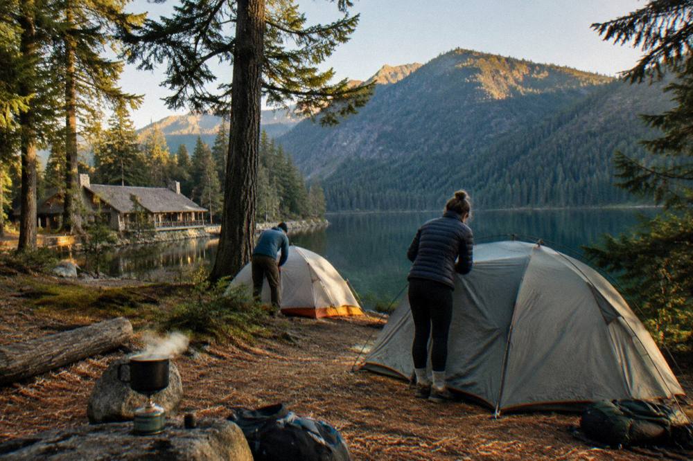 Backpackers setting up tents at a forested lakeside camp beside still water