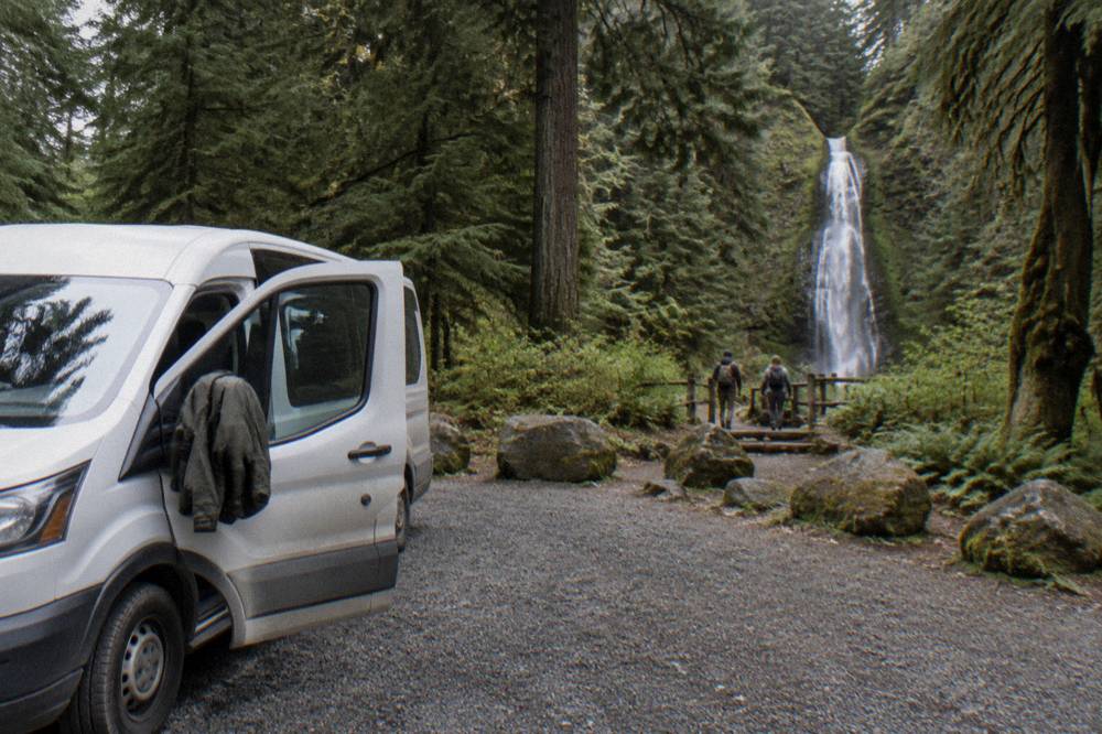A tour van parked at a forest trailhead beside tall evergreens